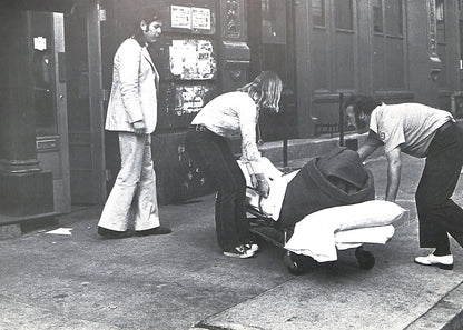 Interior page - CAROLINE TISDALL / JOSEPH BEUYS: COYOTE. Black and white photo of a person being pushed in a wheelchair by two others on a city street.