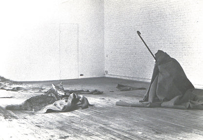 Black and white photograph of a person sitting on the floor with a stick in a minimalistic room.