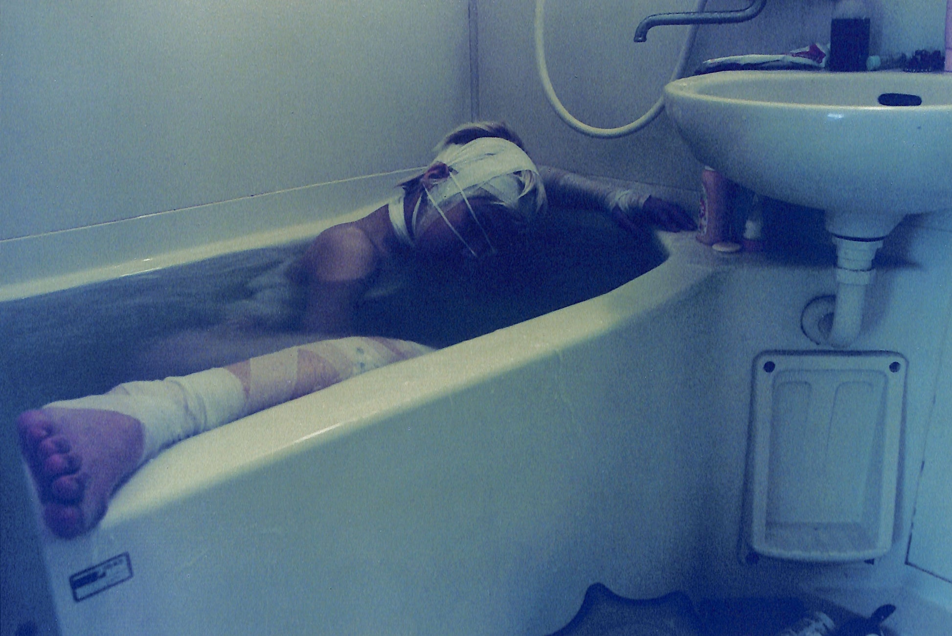 Interior shot - YURIE NAGASHIMA / YURIE NAGASHIMA. Person lying in a bathtub with bandages on, next to a sink.