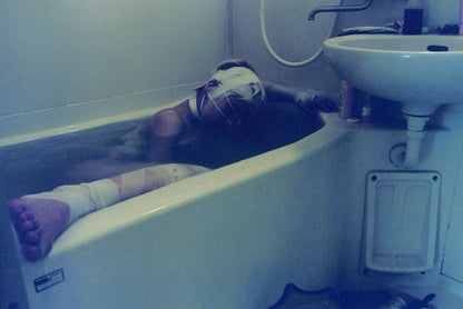 Interior shot - YURIE NAGASHIMA / YURIE NAGASHIMA. Person lying in a bathtub with bandages on, next to a sink.