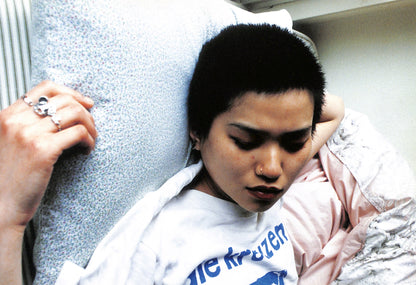 Interior shot - YURIE NAGASHIMA / YURIE NAGASHIMA. Person wearing a white shirt with blue text, sitting on a bed.
